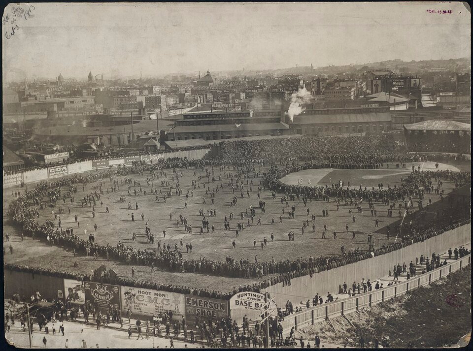 A crowded ballpark photographed from above, thousands of spectators standing on the outfield grass and lining the fences, with a city skyline beyond.