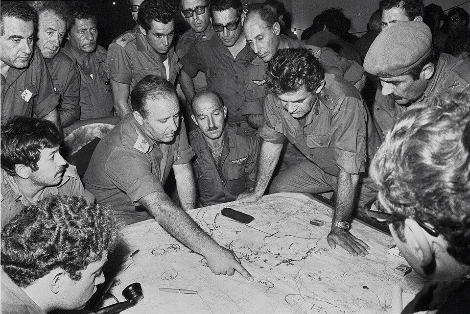 A group of officers in field uniform leaning over a large map spread on a table, one pointing to a marked position, others watching closely.