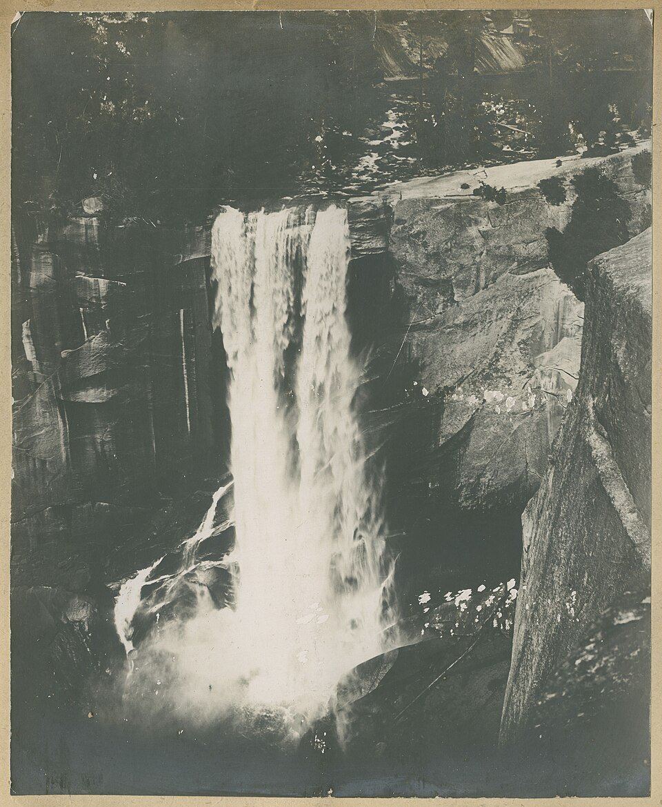 A tall waterfall pouring over a granite ledge into a mist-filled gorge, photographed in sepia with dark forest along the cliff top.